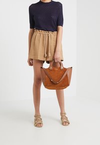 Mujer que lleva una blusa de punto en color navy y pantalones cortos de tiro alto en tono marrón claro, sosteniendo un bolso de cuero y ante marrón, de pie en sandalias de tiras beige.