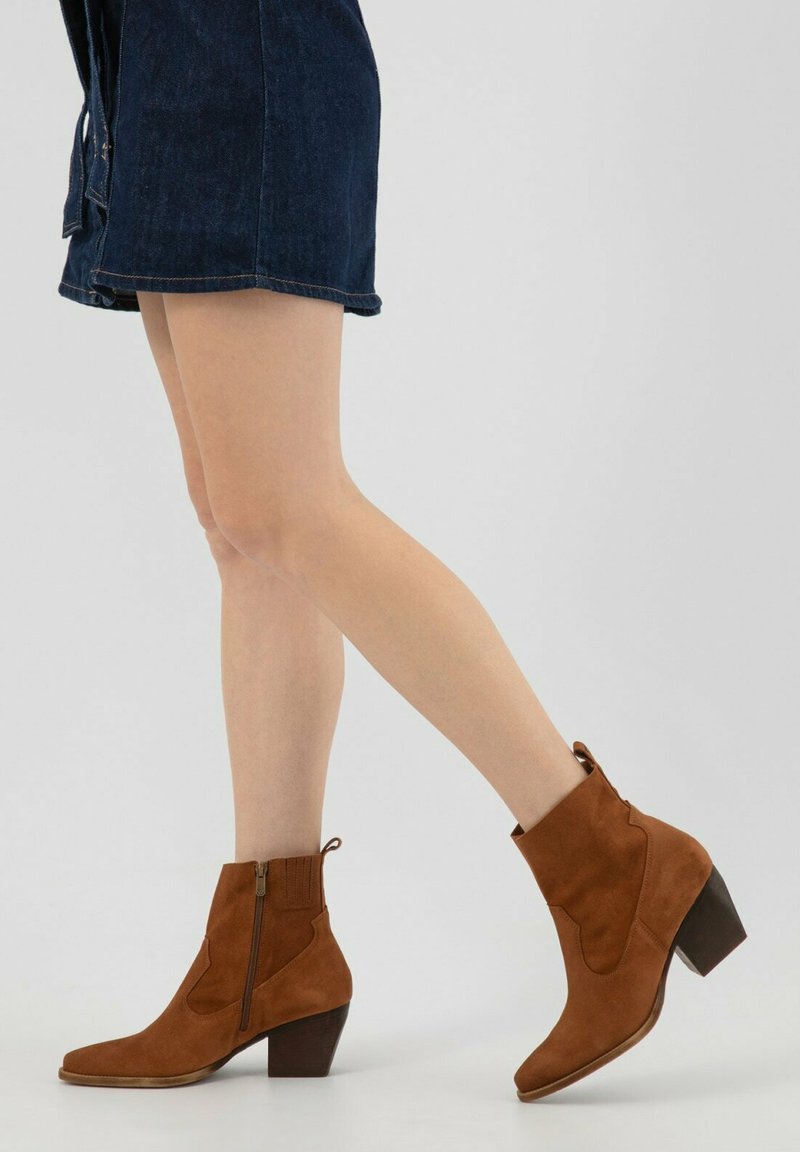 Brązowe zamszowe botki za kostkę z ostrym noskiem, bocznym zamkiem błyskawicznym i wymodelowaną cholewką. Obcas jest krótki i kwadratowy.