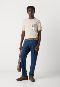 Beige bomull T-skjorte med lomme med grå grafikk, sammen med blå denim jeans, brun belte, olivengrønne sneakers og en grå veske.