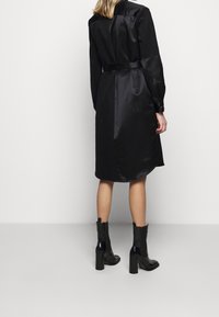 Svart satin klänning med långa ärmar, midja med bälte och en lätt rundad nederdel. Bärs med blanka svarta klackade ankelskor.