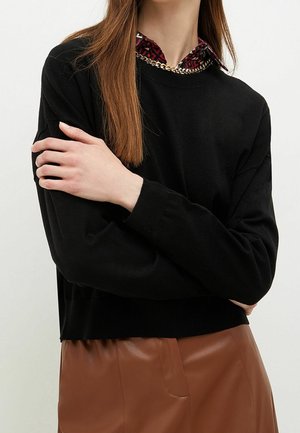 Kvinde med langt, glat hår iført en sort sweater over en rød mønstret skjorte med krave og en brun lædernederdel, armene krydsede.