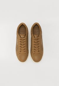Kreem sneakers sile pehmeast nahast varbast ja mustrilistest kangast külgedest. Omavad pruunisid paelu ja valget kummist tallat. Logo on reljeefselt kujutatud keele peal.