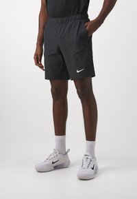 Shorts deportivos negros con cintura elástica, bolsillos laterales y logotipo de la marca Nike en blanco en la pierna izquierda. Combinados con zapatillas blancas.