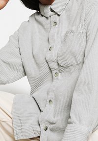 Osoba w jasnoszarej, prążkowanej koszuli z guzikiem i kieszonką na piersi oraz w beżowych spodniach, siedząca na tle białym.