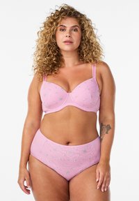 Mujer de cabello rizado con sujetador floral rosa a juego y ropa interior de cintura alta, de pie con un tatuaje en el antebrazo izquierdo.