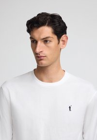 Camicia bianca a maniche lunghe realizzata in cotone, con un piccolo logo blu navy di un golfista sul lato sinistro del petto. Tessuto liscio e scollatura a girocollo.