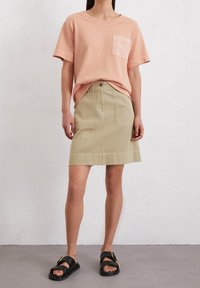 Persikanvärinen puuvillainen t-paita, jossa on rintatasku, yhdistetty beigeen polvipituiseen hameeseen, jossa on etutaskut ja metalliset nappisulkimet. Mustat sandaalit täydentävät asun.