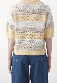 Pletený pulovr v krémové, béžové a žluté barvě s otevřenými vzory ve tvaru diamantu; krátké rukávy a žebrovaný límec. Nosený přes džíny s vysokým pasem.