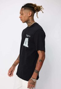Personne avec des dreadlocks et des tatouages portant un t-shirt noir avec un graphique de l'Arc de Triomphe et un pantalon blanc, se tenant contre un fond uni.