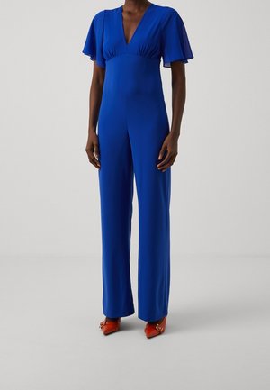 Persoon staat en draagt een blauwe jumpsuit met volantmouwen en oranje puntige hakken met gespdetails.