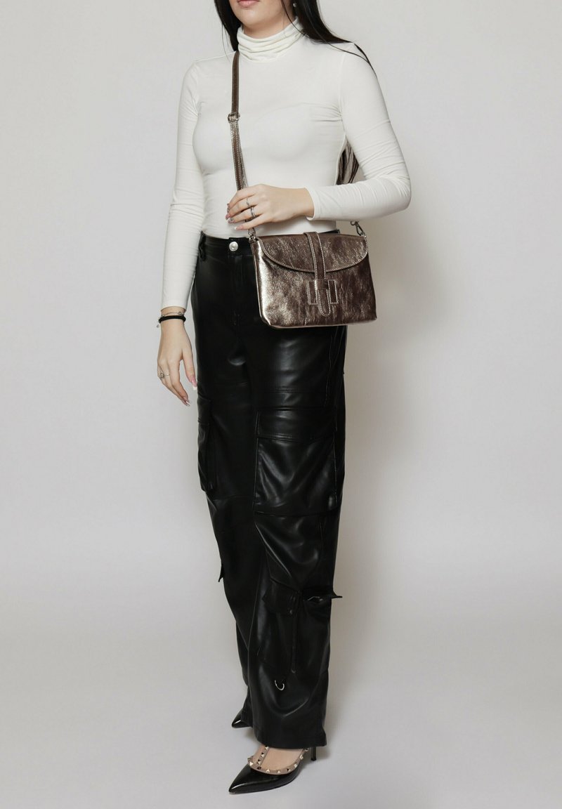 Borsa in bronzo metallico con chiusura a pattina e dettagli decorativi a strisce verticali. Indossata con un dolcevita bianco e pantaloni cargo in pelle nera.