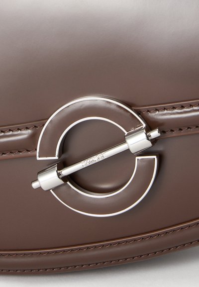 Gros plan d'un fermoir circulaire en argent avec une barre horizontale gravée de "SAND COPENHAGEN" sur un cuir marron foncé cousu.