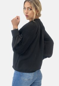 Svart plisserad blus med långa puffärmar, har en rundad halslinning och mjukt, slätt tyg. Bärs över ljusblå jeans.