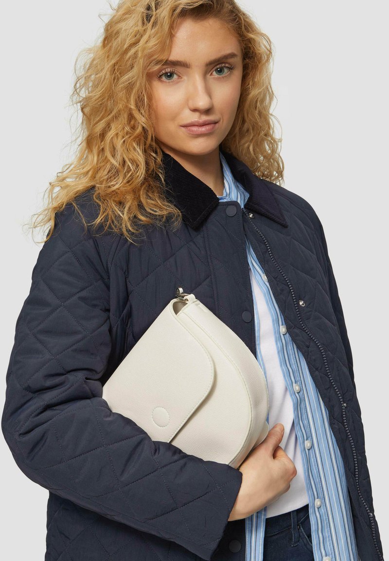 Marineblau gesteppte Jacke mit einem Cordkragen, cremefarbene Clutch mit abgerundetem Klappenverschluss, über einem hellblau gestreiften Hemd getragen.