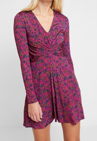 Sukienka z długim rękawem i dekoltem w kształcie V w różowo-granatowy kwiatowy wzór, z detalem w postaci związanej talii oraz zwiewnym dołem. Gładka tekstura materiału.