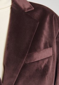 Gros plan sur le revers d'un blazer en velours marron foncé et une poche poitrine, porté sur un haut en maille côtelée crème.