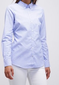 Camicia azzurra a maniche lunghe con colletto, caratterizzata da bottoni bianchi, una texture liscia e un orlo curvo. Indossata con pantaloni bianchi.