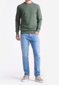 Man in een olijfgroen sweatshirt, lichte blauwe jeans, beige sneakers, staand met één hand in de zak op een witte achtergrond.