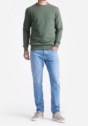 Man in een olijfgroen sweatshirt, lichte blauwe jeans, beige sneakers, staand met één hand in de zak op een witte achtergrond.