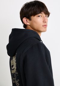 Black hoodie with a drawstring, featuring a graphic design on the back. Material appears soft; the design includes circular patterns and text.