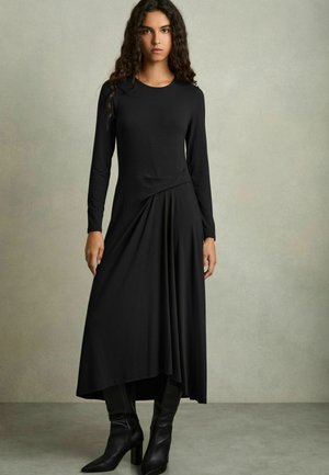 Black long-sleeve dress with a round neckline, draped design at the waist, and asymmetrical hem; paired with high-heeled black boots.