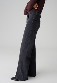 Jean en denim noir à jambe large avec taille haute, présentant une texture subtile et un design à cinq poches. Fini avec des coutures contrastées.