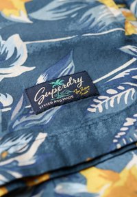 Tessuto floreale blu navy con accenti gialli, caratterizzato da grandi foglie e fiori. Include un'etichetta tessuta nera con "Superdry" e logo.