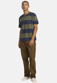 Striped cotton t-shirt in navy, olive green, and beige, featuring a chest pocket and a relaxed fit, paired with brown pants and casual sneakers.