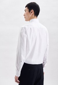 Camicia bianca con colletto a fascia e maniche lunghe, dotata di polsini con bottoni. Indossata con pantaloni scuri, mostrando una texture di tessuto liscia.