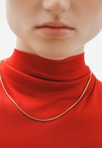 Collarín de cadena de oro descansando sobre una blusa de tela roja de cuello alto. Textura suave, brillo sutil y diseño minimalista enfatizado por el contraste de colores.
