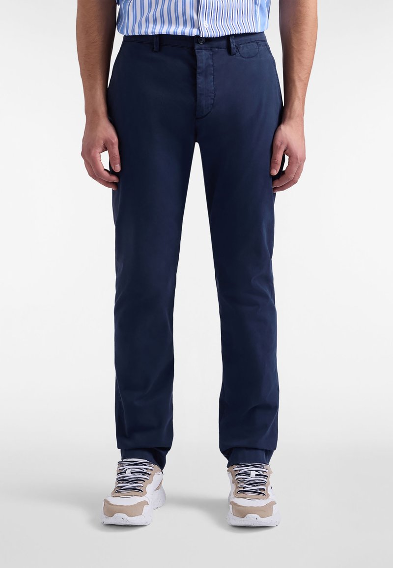 Pantaloni di cotone blu navy con taglio dritto, dotati di passanti per cintura e cerniera frontale. Indossati con sneakers beige e bianche.