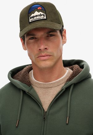 Hombre con gorra de pana verde oliva con logo de montaña y sudadera con cremallera verde con forro de piel marrón sobre suéter beige.