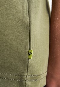 Camiseta verde oliva con una textura suave, que presenta una etiqueta amarilla contrastante con un logo negro en la costura lateral y detalles de costura cuidados.