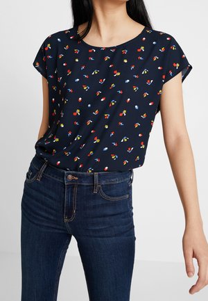 Woman wearing a navy short-sleeve blouse with colorful abstract shapes paired with dark blue high-waisted denim jeans.