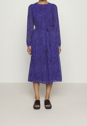 Kobieta w fioletowej sukience midi z czarnymi abstrakcyjnymi liściastymi wzorami, z wcięciem w talii, długimi rękawami i czarnymi sandałami na obcasie na odkryte palce.