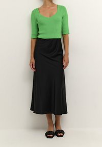 Grøn ribbet strikbluse med v-udskæring, sort satin midi-nederdel og sorte flettede slidesandaler. Glatte teksturer og tætsiddende silhuet.