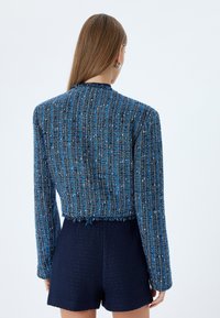 Blå tweed cropped jakke med tekstureret stof og frynsekanter, parret med mørkeblå teksturerede shorts. Simpelt design uden udsmykninger.