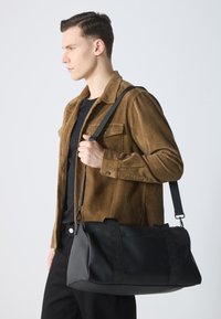 Sac de sport noir en tissu robuste, de forme rectangulaire, avec deux anses supérieures. Il dispose d'une longue sangle réglable et d'un design minimaliste.