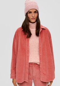 Veste en velours côtelé rose associée à un pull col roulé en tricot rose clair et un pantalon assorti, complétée par un bonnet rayé rose.