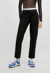 Pantalons noirs à coupe droite, dotés de passants pour ceinture, de poches latérales et d'une texture lisse, associés à des chaussures de sport rehaussées de bleu.