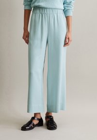 Vaaleansinisiä, leveälahkeisia housuja, jotka on valmistettu sileästä kankaasta ja joissa on joustava vyötärö. Yhdistettynä mustiin, hihnallisiin sandaaleihin, joissa on solkiyksityiskohta.