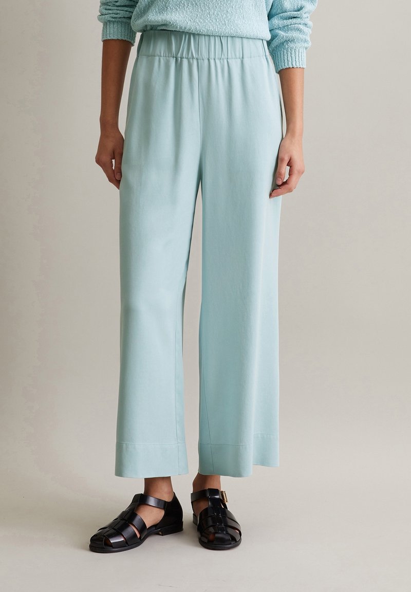 Vaaleansinisiä, leveälahkeisia housuja, jotka on valmistettu sileästä kankaasta ja joissa on joustava vyötärö. Yhdistettynä mustiin, hihnallisiin sandaaleihin, joissa on solkiyksityiskohta.