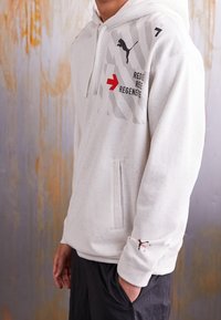 Bílá mikina s kapucí s tečkovanou texturou, která má šedý vzor, červené grafické detaily a černé logo na hrudi a rukávu. Součástí je klokaní kapsa.