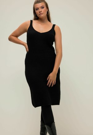 Zwarte gebreide jurk met een V-hals en brede bandjes, aansluitende silhouet en een jurk tot de knie, gecombineerd met zwarte leggings en cowboylaarzen.