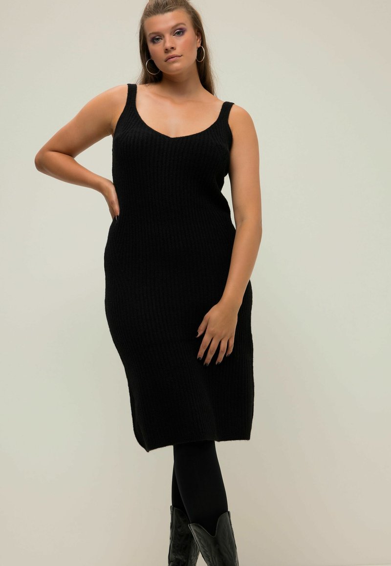 Abito nero a maglia con scollatura a V e spalline larghe, silhouette aderente e orlo fino al ginocchio, abbinato a leggings neri e stivali da cowboy.