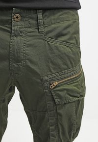Pantaloni cargo verde oliva realizzati in materiale leggero. Dotati di tasche con zip e tessuto testurizzato con cuciture tonali. Design casual.