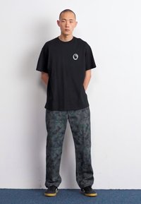 Camiseta de algodón negra con un pequeño logo en el pecho, combinada con pantalones de camuflaje grises. El calzado es oscuro con suelas claras.