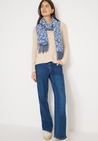 Écharpe à imprimé léopard bleu avec franges, portée sur un pull côtelé beige et associée à un jean évasé bleu. Texture douce, design décontracté.