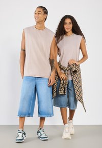 Dva posameznika nosita svetlo bež brezrokavnike. Moški ima na sebi prevelike svetlo modre denim kratke hlače in športne copate, medtem ko ima ženska okoli pasu uvoženo karirano srajco.