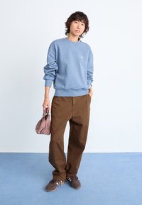 Light blue sweatshirt, brown wide-leg pants, and brown sneakers with pink accents. Carrying a small, pink pouch bag. Smooth textures.